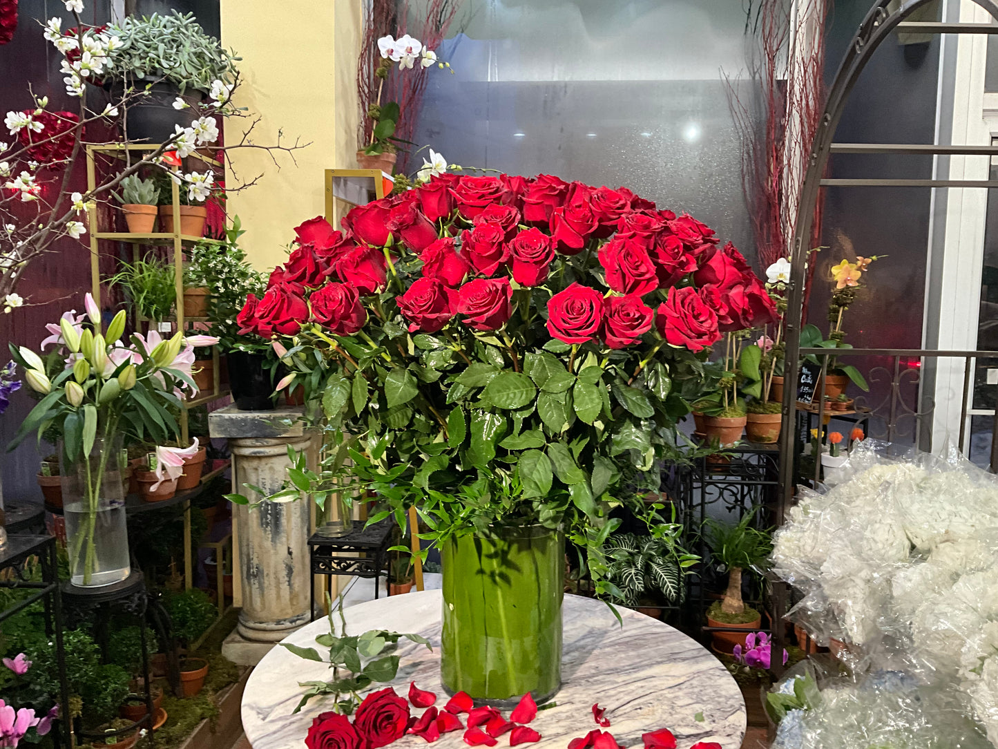 Complete Love small size: one tall glass vase of long-stem red roses on a marble table with scattered petals