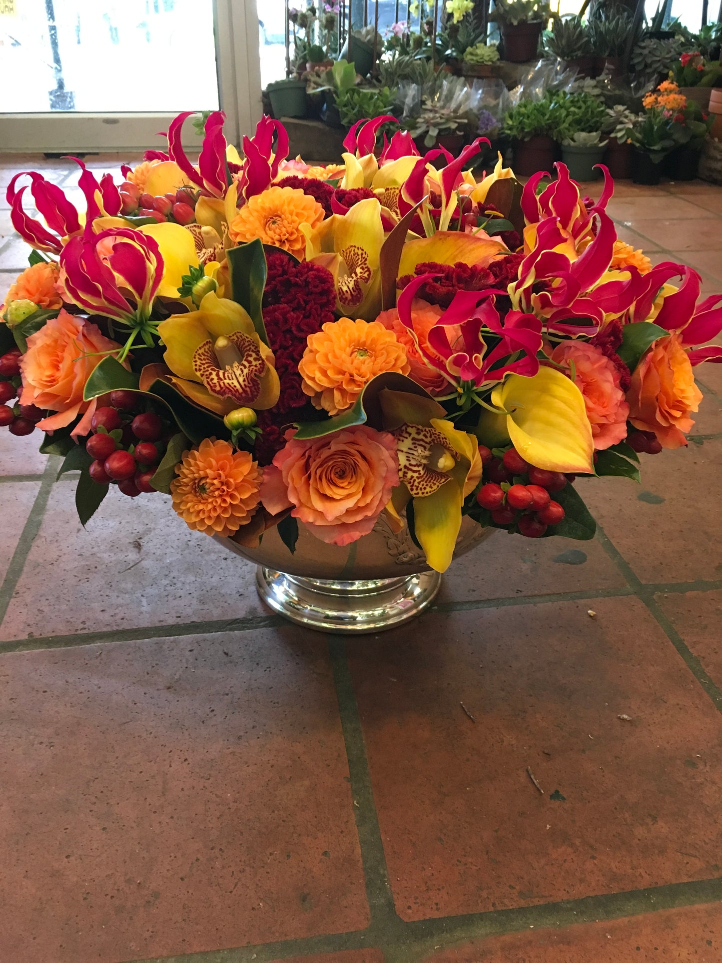 Red Berry centerpiece of orange roses, gloriosa lilies, yellow cymbidium orchids and red berries in a silver footed bowl