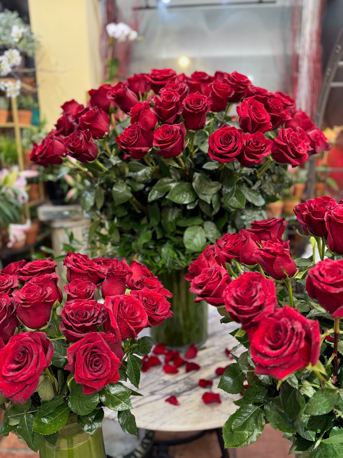 Complete Love, close view of a tall red rose arrangement with rose petals on a marble table and red roses in front