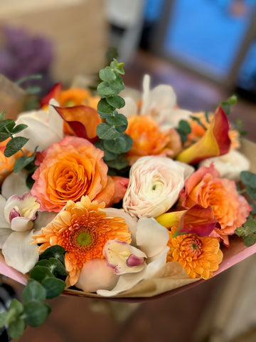 Erly Riser close-up of an orange rose, cream ranunculus, orange gerbera and a white cymbidium orchid with red-orange callas