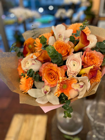 Erly Riser hand-tied in kraft paper with pink tissue: orange roses and gerberas, white orchids, small dahlias and eucalyptus