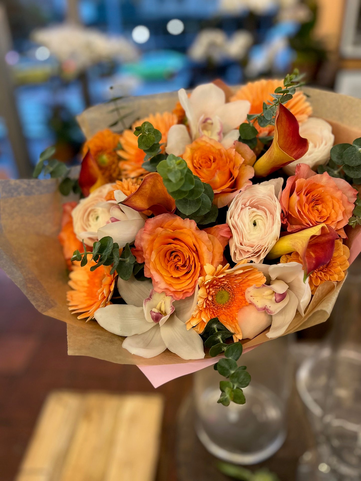 Erly Riser hand-tied in kraft paper with pink tissue: orange roses and gerberas, white orchids, small dahlias and eucalyptus