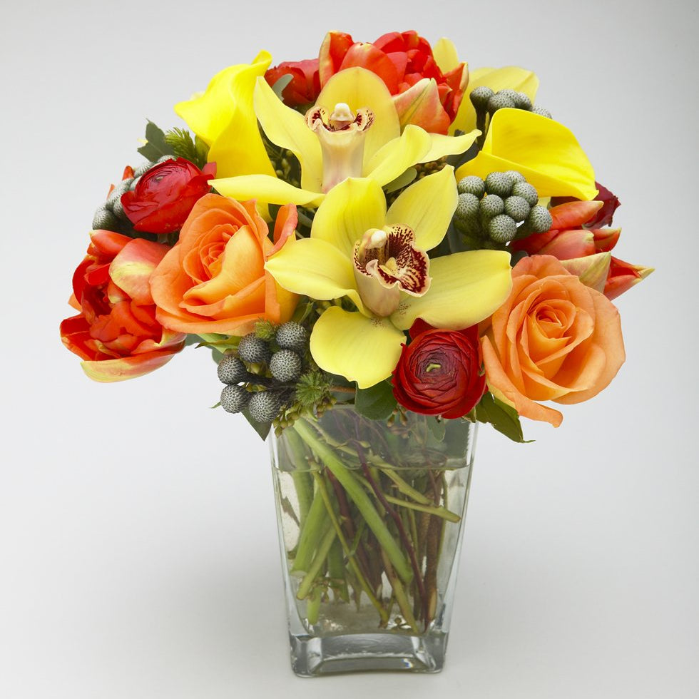 Enticement: yellow cymbidium orchids and calla lilies, orange roses, orange-red tulips and red ranunculus in a glass vase
