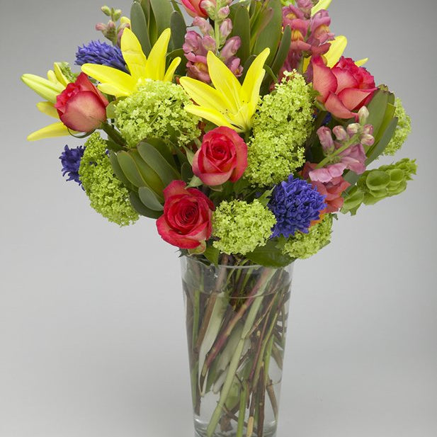 Kaleidoscope yellow lilies, pink roses, pink snapdragons, green viburnum and blue hyacinth in a tall clear glass vase