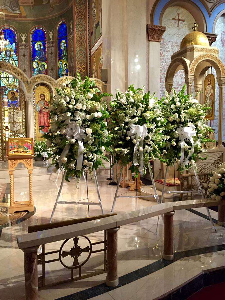 Eternal Rest sympathy sprays, three shown, of white roses, lilies and green hydrangea on easels at a church altar