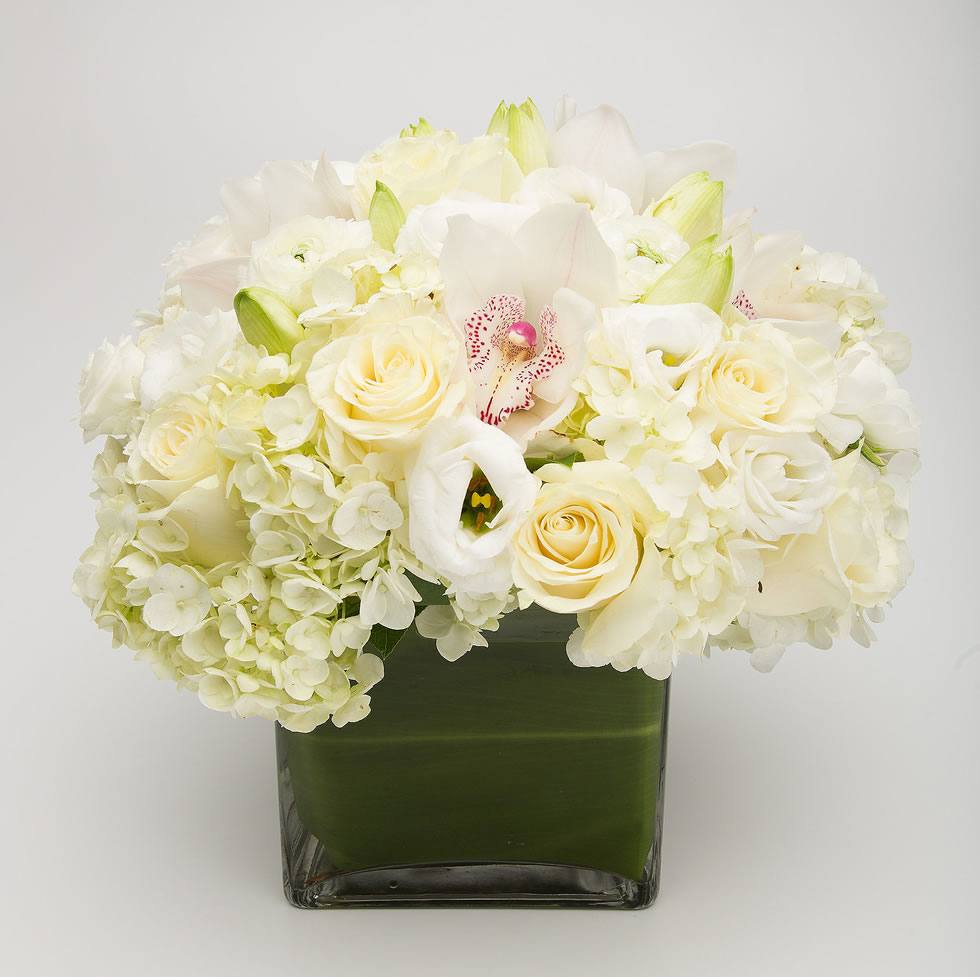 A floral arrangement featuring white hydrangea, cymbidium orchid, roses, lusianthus, and lilies in a clear glass cube vase.