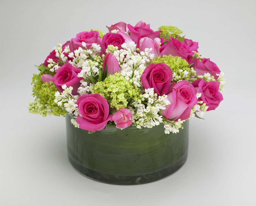 Sweetheart, hot pink roses, pink tulips, white lilac and green viburnum in a low round leaf-lined glass vase