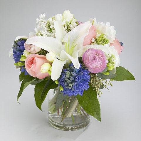 Lullaby arrangement with a white lily, pink roses, pink ranunculus, blue hyacinth and white freesia in a cylinder vase