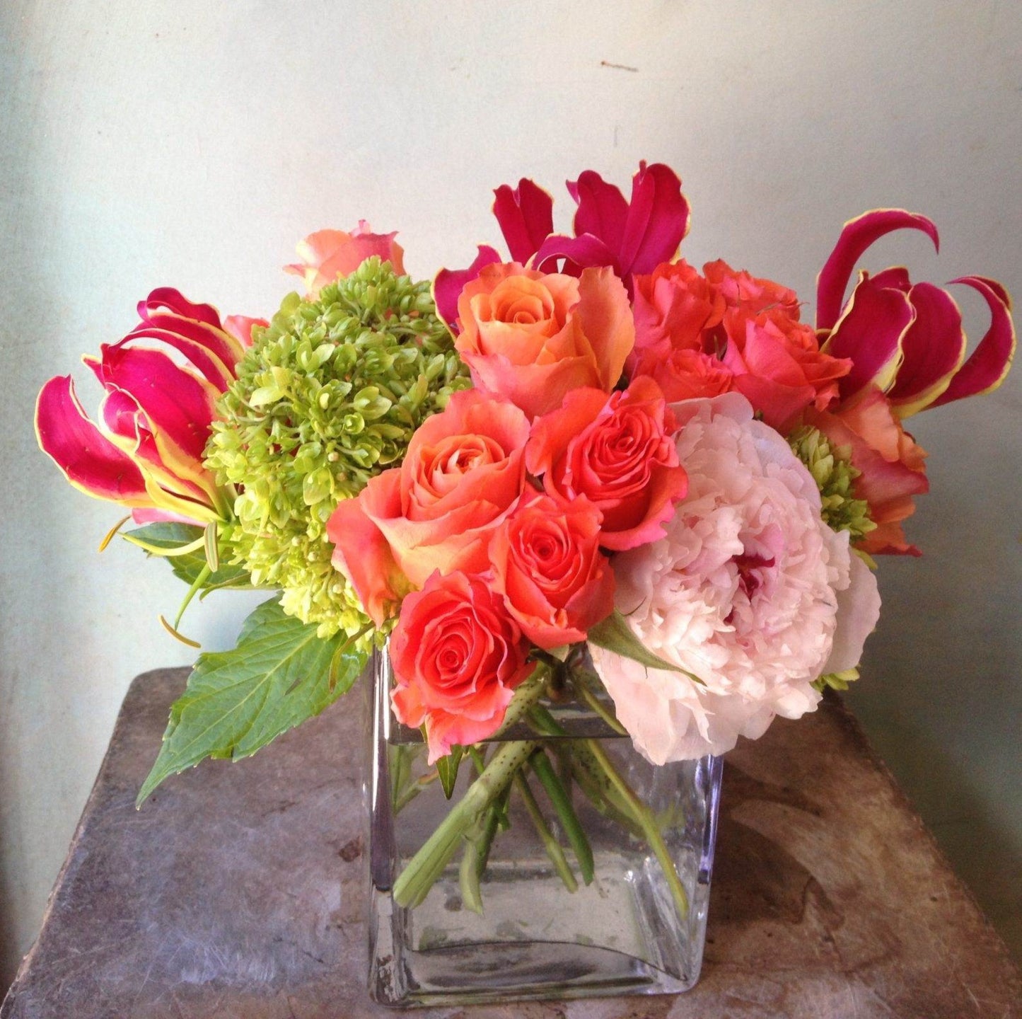 Summer Sunset, orange roses, hot pink gloriosa lilies, a blush peony and green hydrangea in a clear glass cube vase