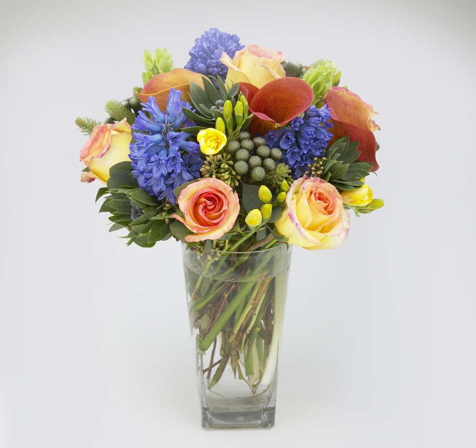 Blue hyacinth, peach roses, rust calla lilies and yellow freesia in a glass vase