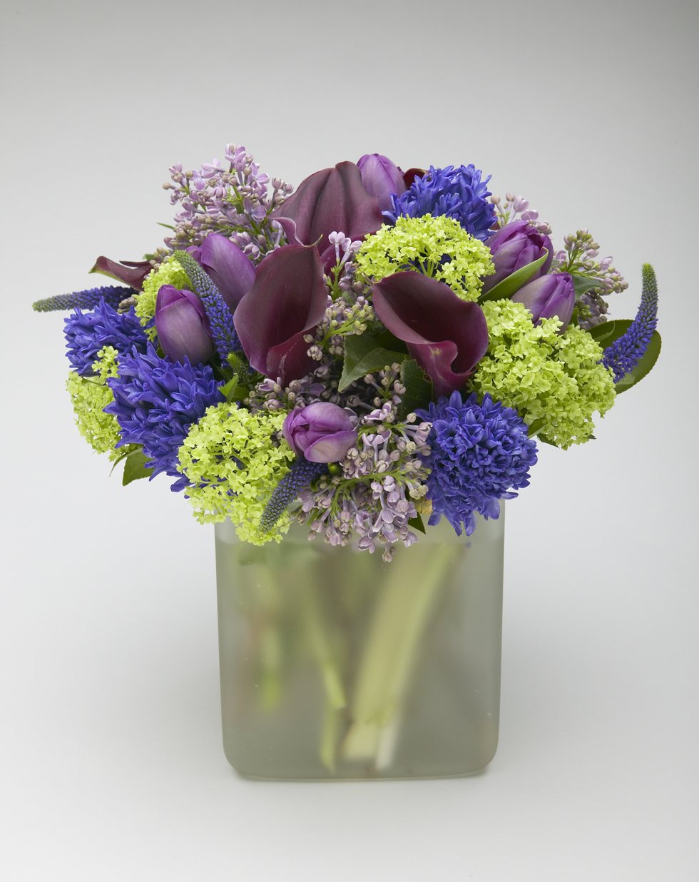 Purple lilac with plum callas, purple tulips, blue hyacinth and green viburnum in a frosted glass vase