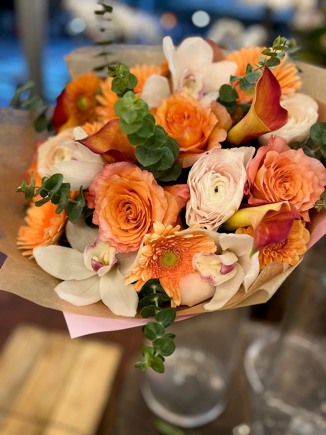 Hand-tied bouquet of orange roses, gerberas, callas, ranunculus and white orchids with eucalyptus, wrapped in kraft paper