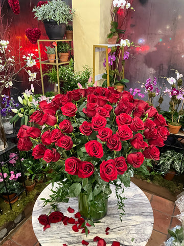 
                    
                      Complete Love: a tall vase of red roses on a marble table with loose roses and petals, orchid plants behind
                    
                  
