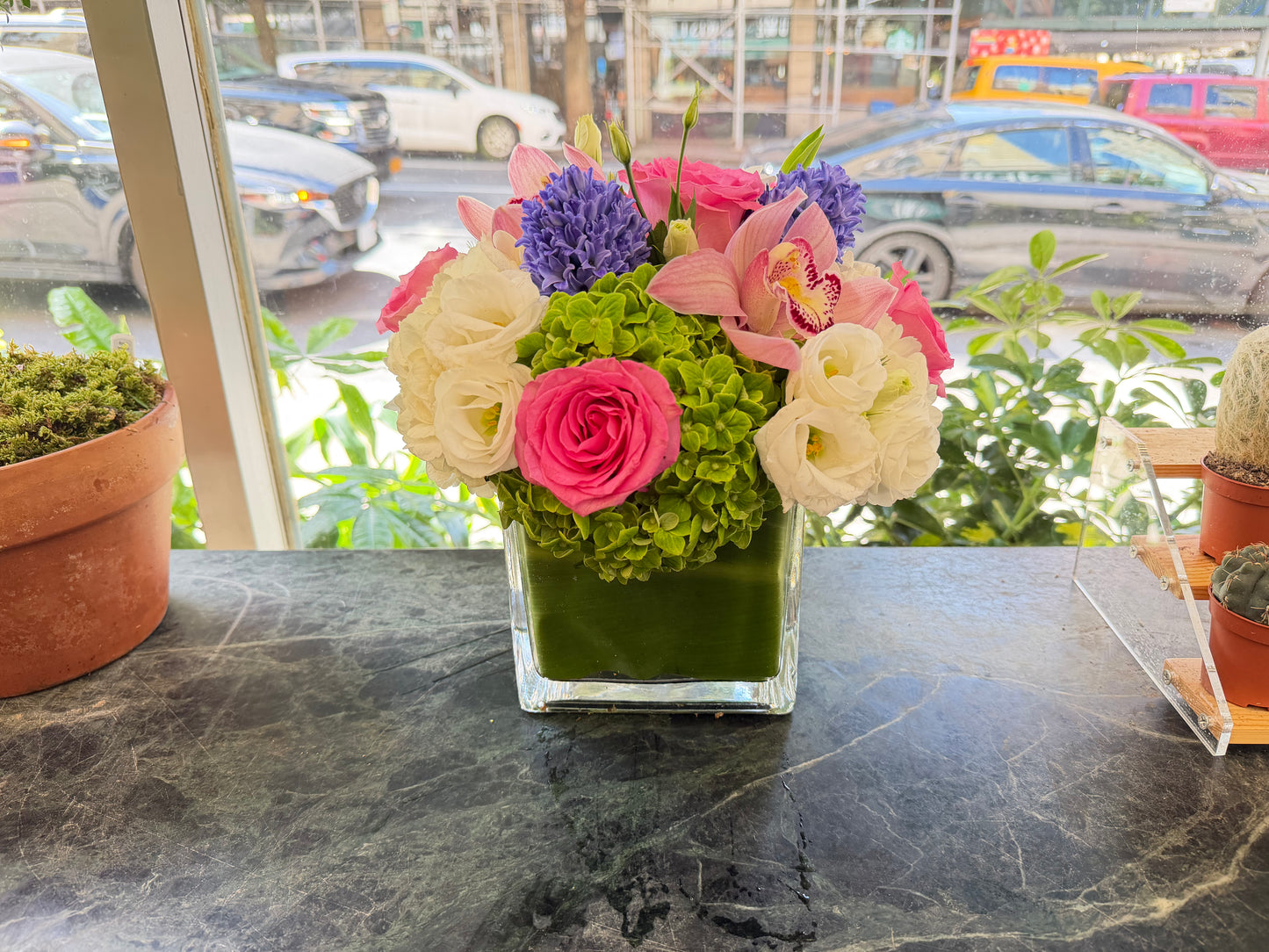
                  
                    Eliana seen from slightly above, hot pink rose front and center, pale pink orchids and purple hyacinth, sunlit street behind
                  
                