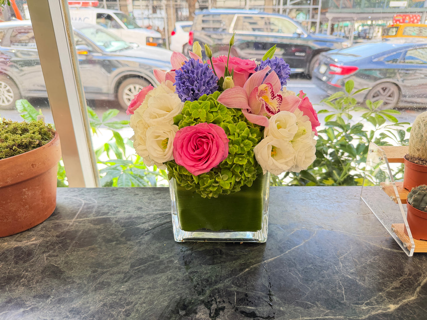 
                  
                    Front view of Eliana with hot pink roses, white lisianthus, blush cymbidium orchids and purple hyacinth on green marble
                  
                