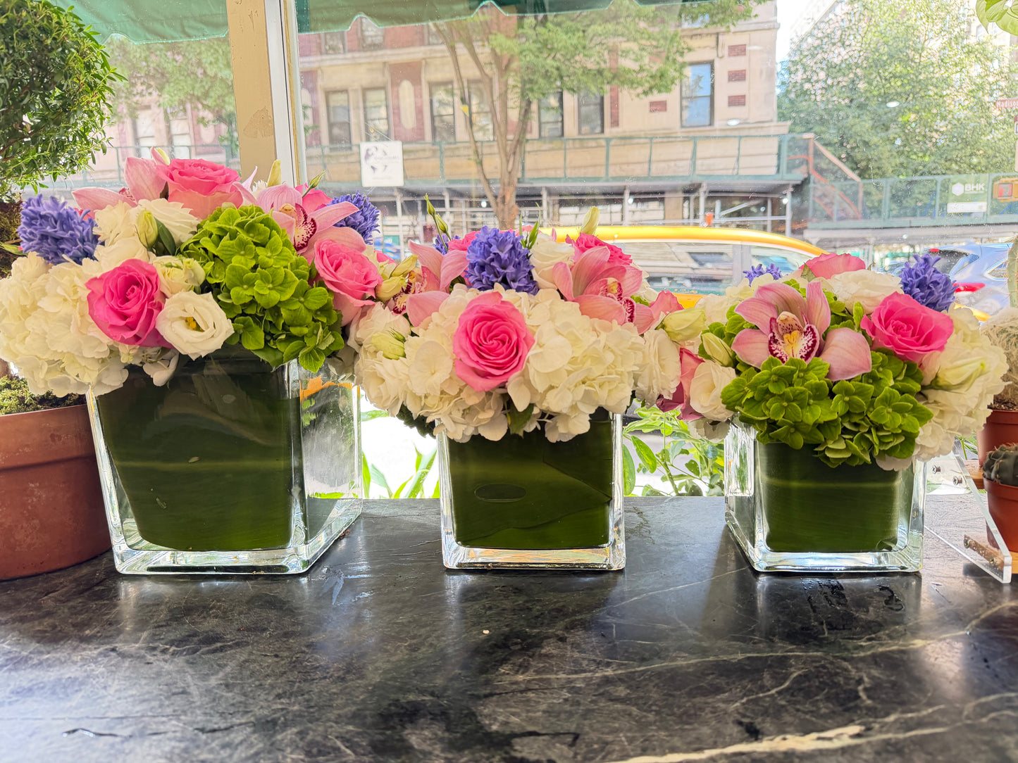
                  
                    Three matching Eliana arrangements on the Jerome Florists front counter with a sunny Madison Avenue street view behind
                  
                