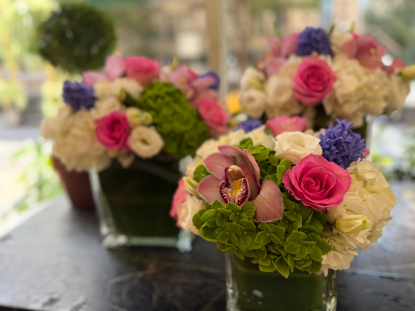Three Eliana arrangements on a dark stone table, bright blurred street behind, front cube sharp with hot pink roses