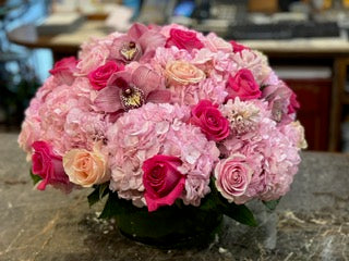 
                    
                      Jab, small size: pink hydrangea with hot pink roses, pink cymbidium orchids and a peach rose in a low dark vase on a counter
                    
                  