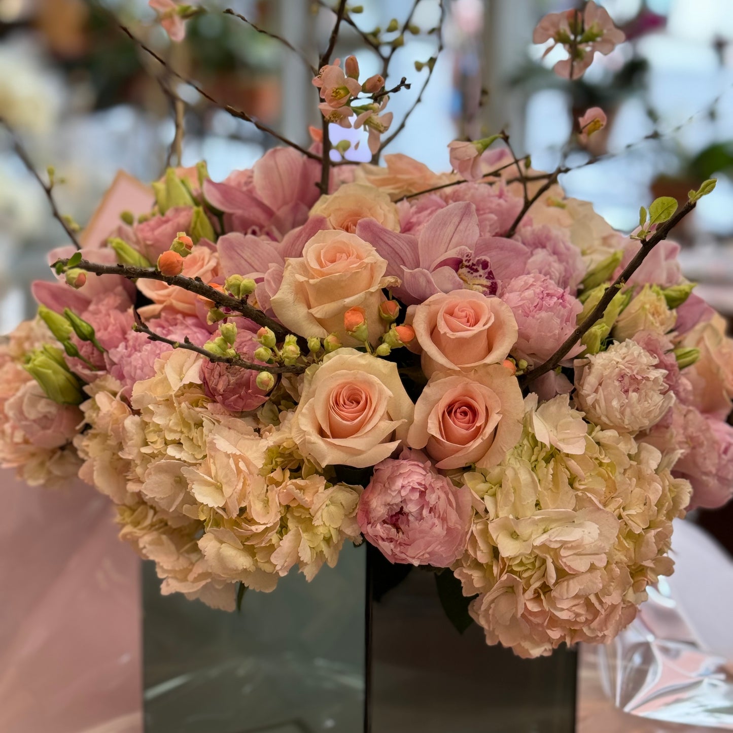 Pink Fury with peach roses, pink peonies, cymbidium orchids and cream hydrangea under blossom branches in a mirrored cube