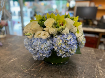 
                    
                      Blue Dream in a round leaf-lined glass bowl: white roses, lisianthus and green cymbidium orchids over blue hydrangea
                    
                  