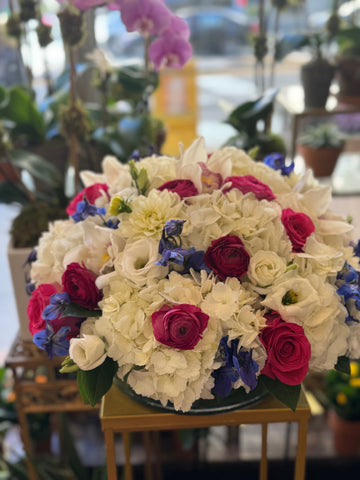 
                    
                      White Fire round centerpiece of white hydrangea, pink roses, ranunculus and blue delphinium on a gold stand in the shop
                    
                  