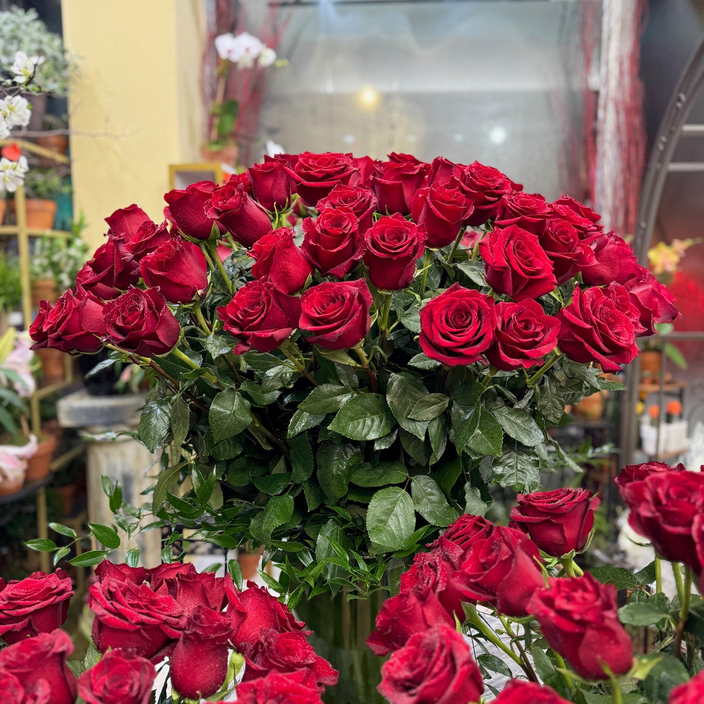 Complete Love: a tall vase of dozens of red roses with more red rose arrangements and petals in the foreground