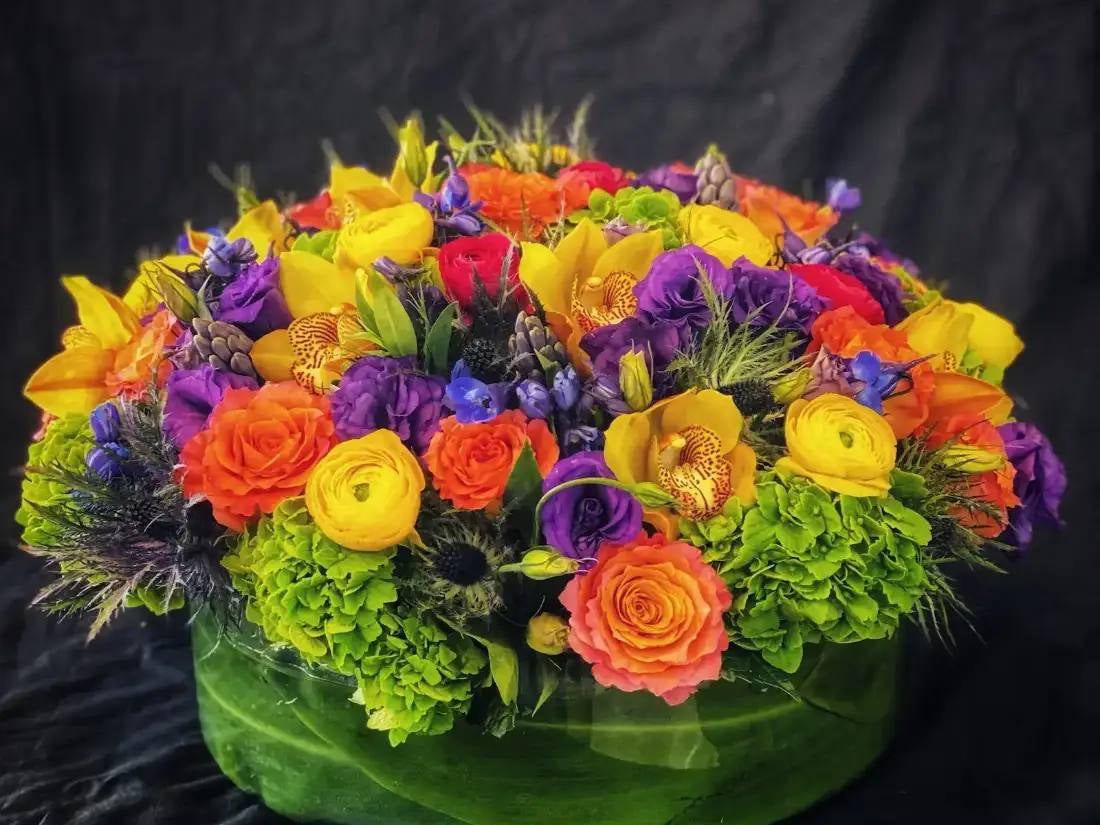 Colorful arrangement of orange roses, yellow ranunculus, orchids, and purple lisianthus in a leaf-wrapped base