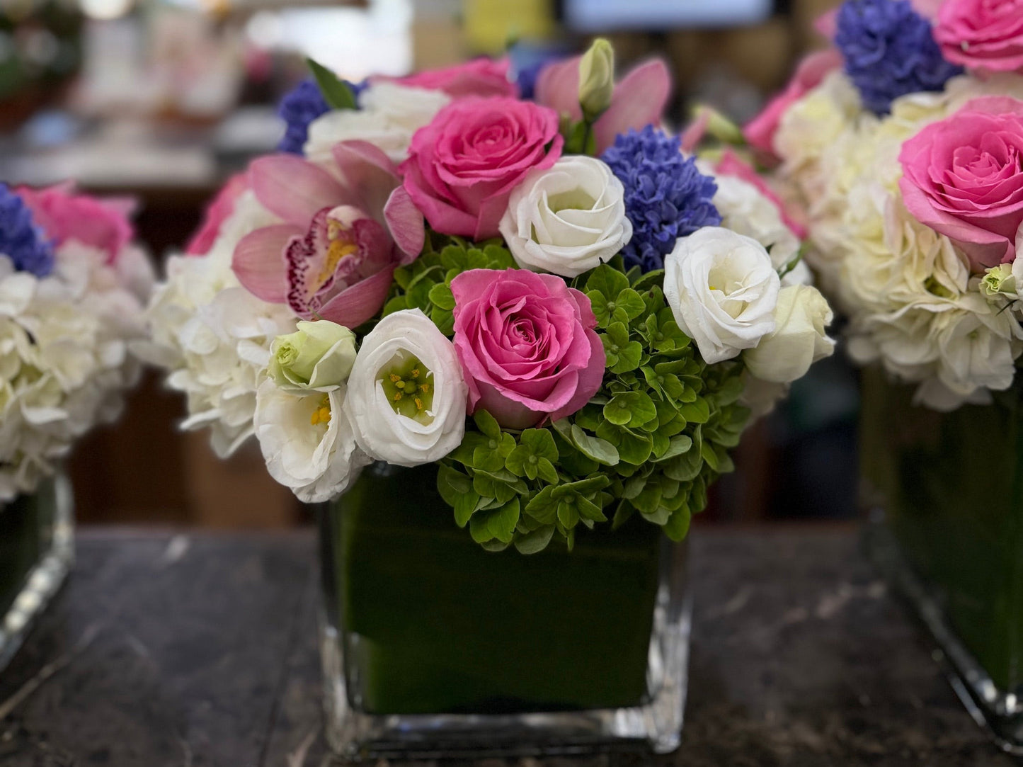 
                  
                    Eliana on the shop counter: pink roses, white lisianthus, green hydrangea and lavender-blue hyacinth in a leaf-lined glass cube
                  
                