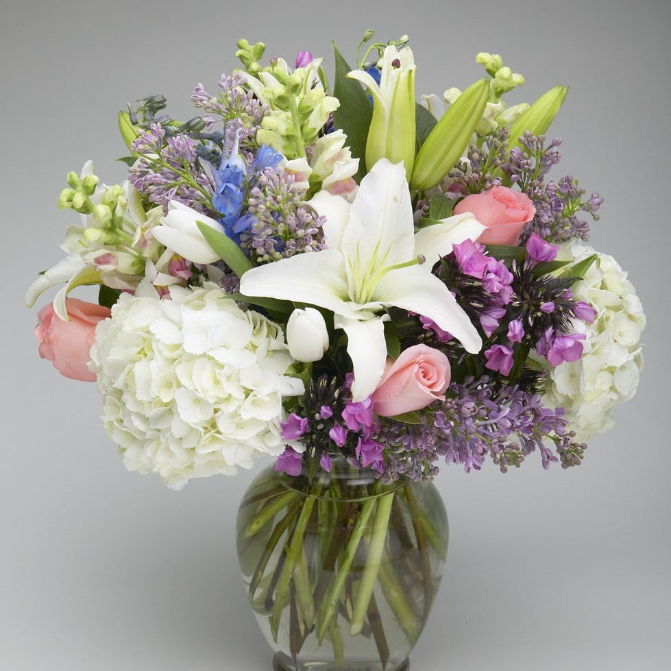 Cinderella pastel arrangement of white lilies, pink roses, white hydrangea, purple lilac and phlox in a clear glass vase
