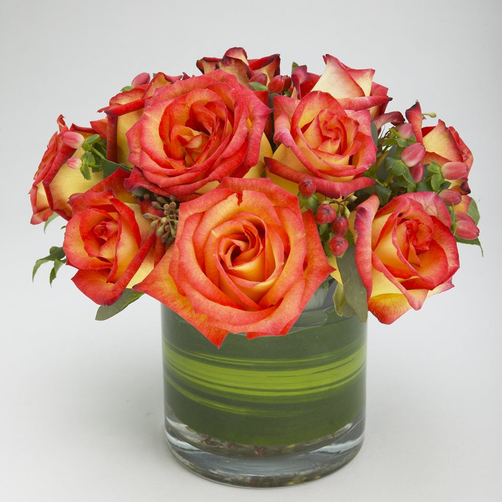Fireball: yellow roses edged in coral red with pink hypericum berries in a leaf-lined glass cylinder vase